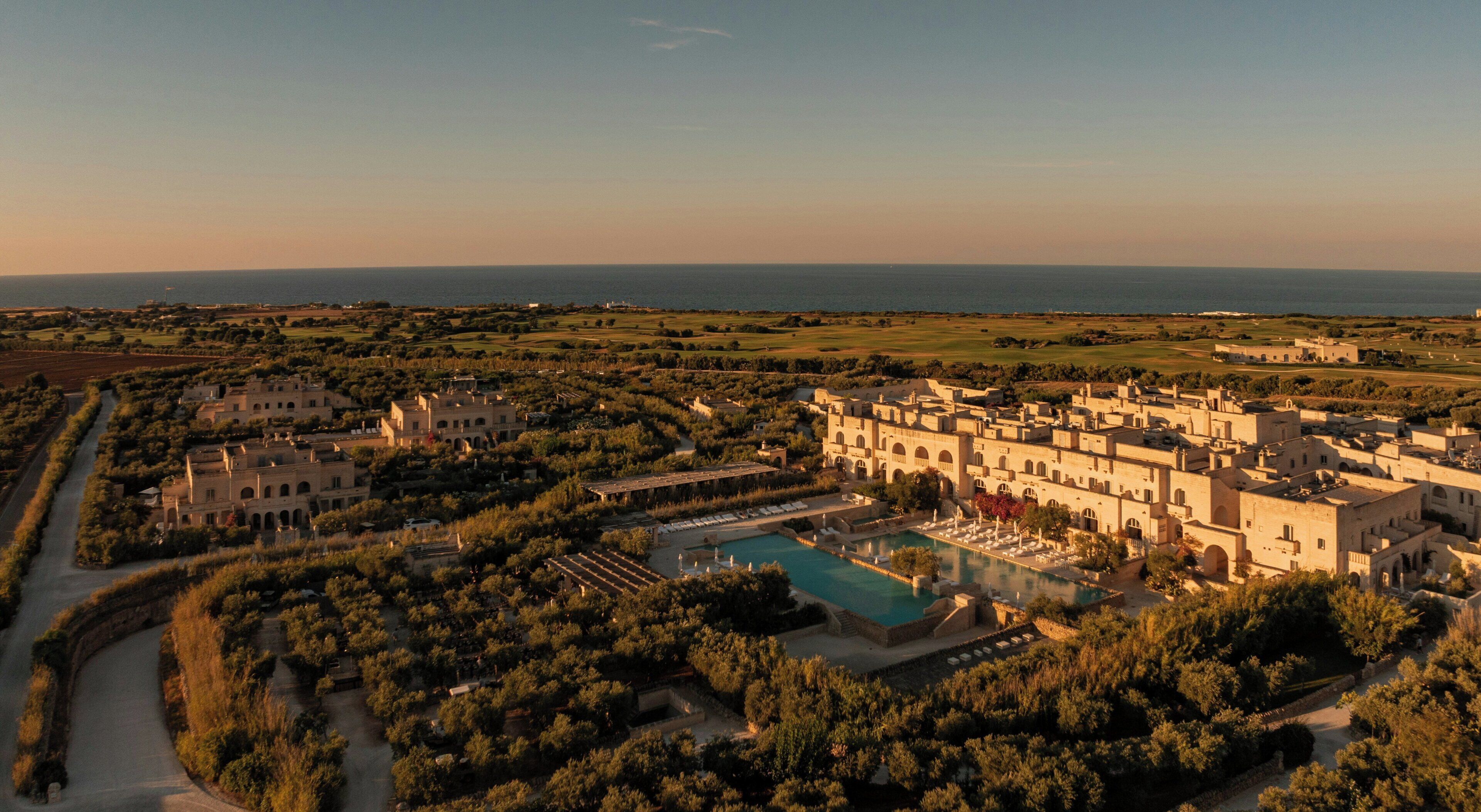 A large resort with multiple buildings, a swimming pool, and a view of the sea.