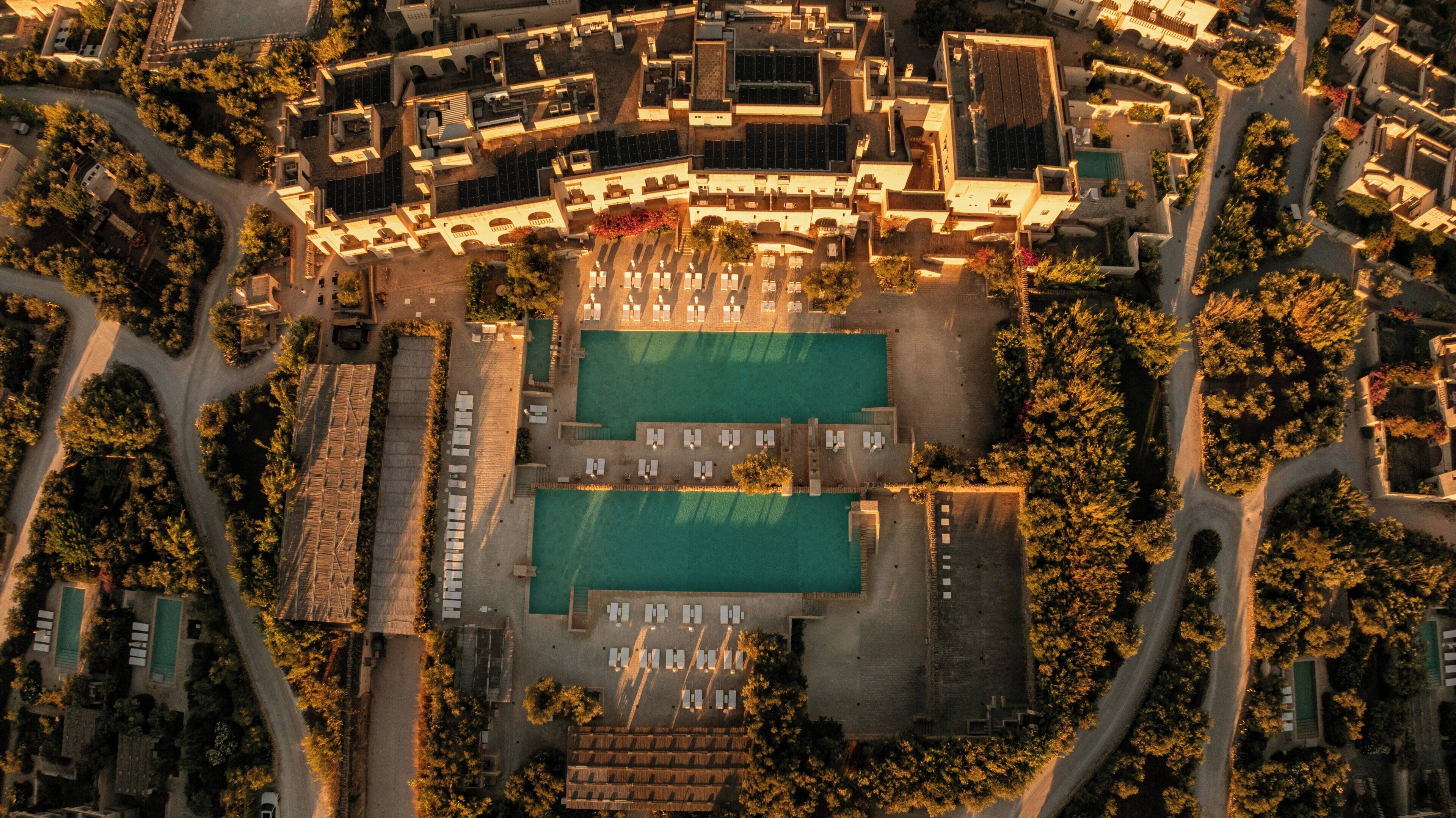 An aerial view of a resort with a central pool area, multiple buildings, and surrounding greenery.