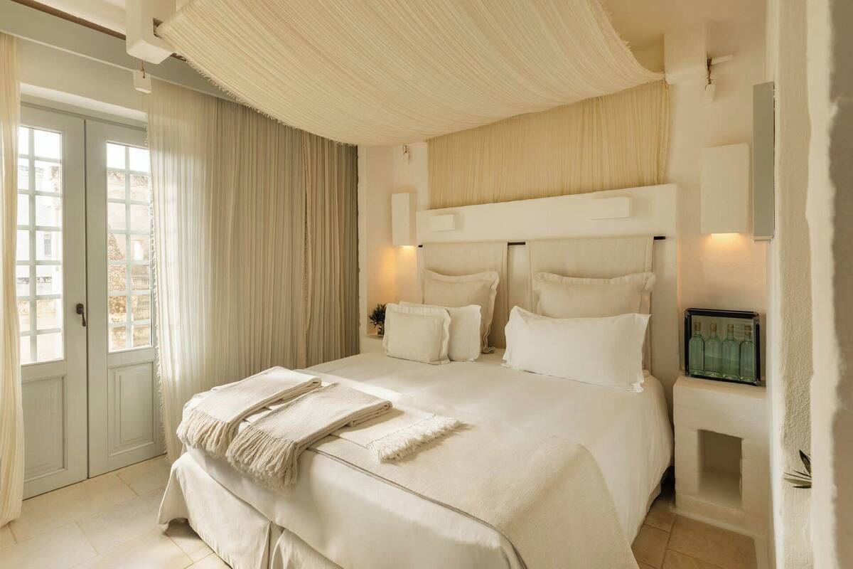 A spacious bedroom with a large bed, a view of the city through the window, and a nightstand with a bottle of water.