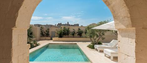 Une piscine avec vue sur le paysage, encadrée par une arche, et entourée de chaises longues et de verdure.