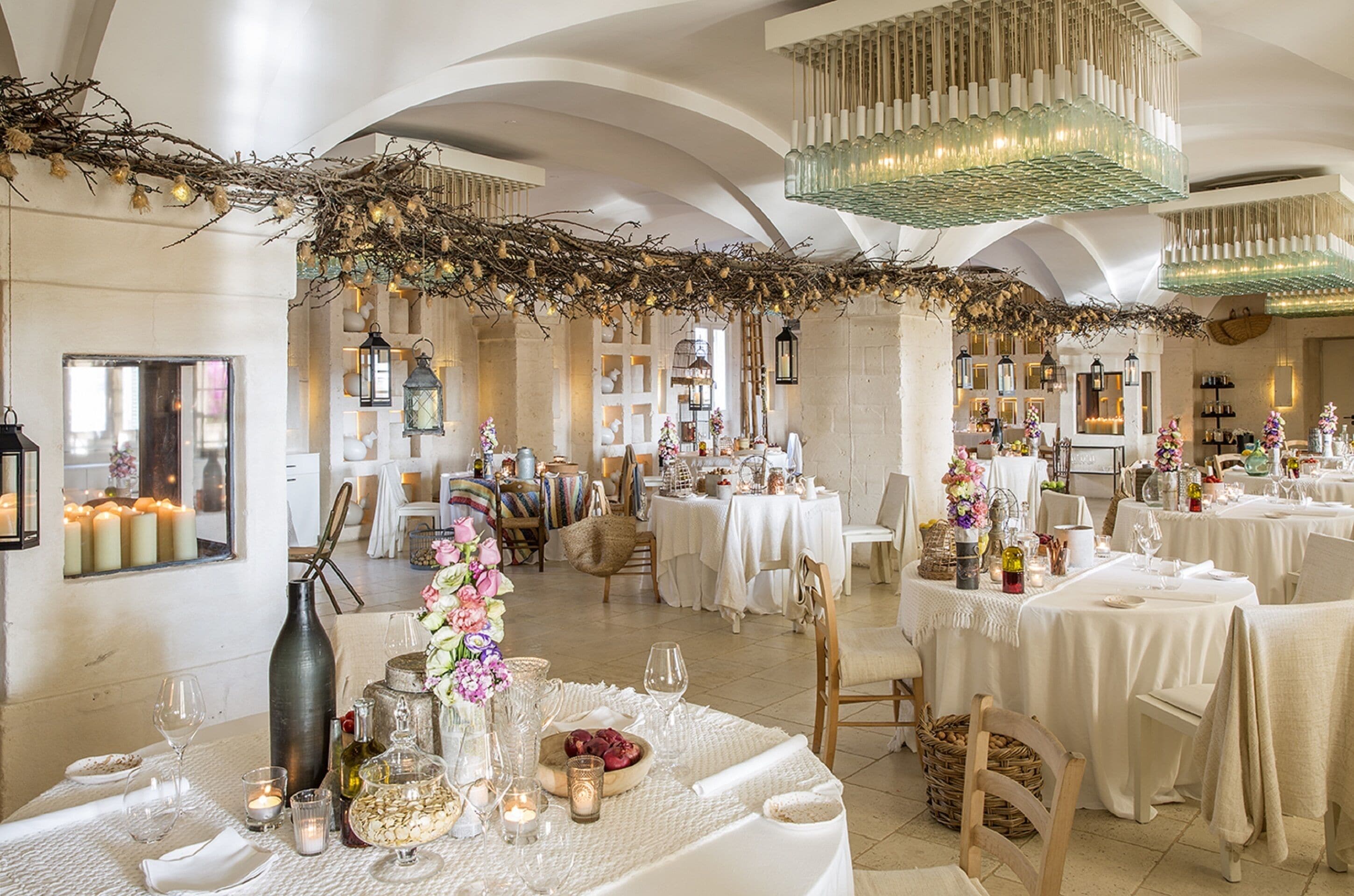 A spacious dining area with multiple tables set for a meal, featuring white tablecloths, candles, and floral centerpieces.