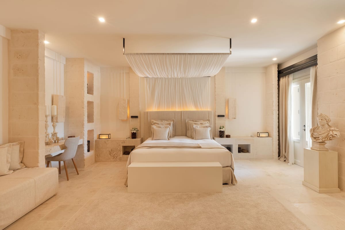 A spacious bedroom with a large bed, bedside tables, a chair, and a sculpture on a pedestal.