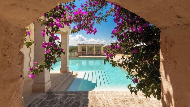 An archway leading to a pool area with a pergola and a view of a building in the background. - Borgo Egnazia (Fasano)