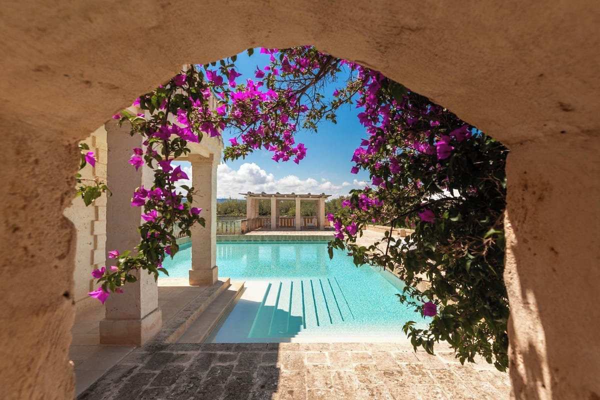 An archway leading to a pool area with a pergola and a view of a building in the background.