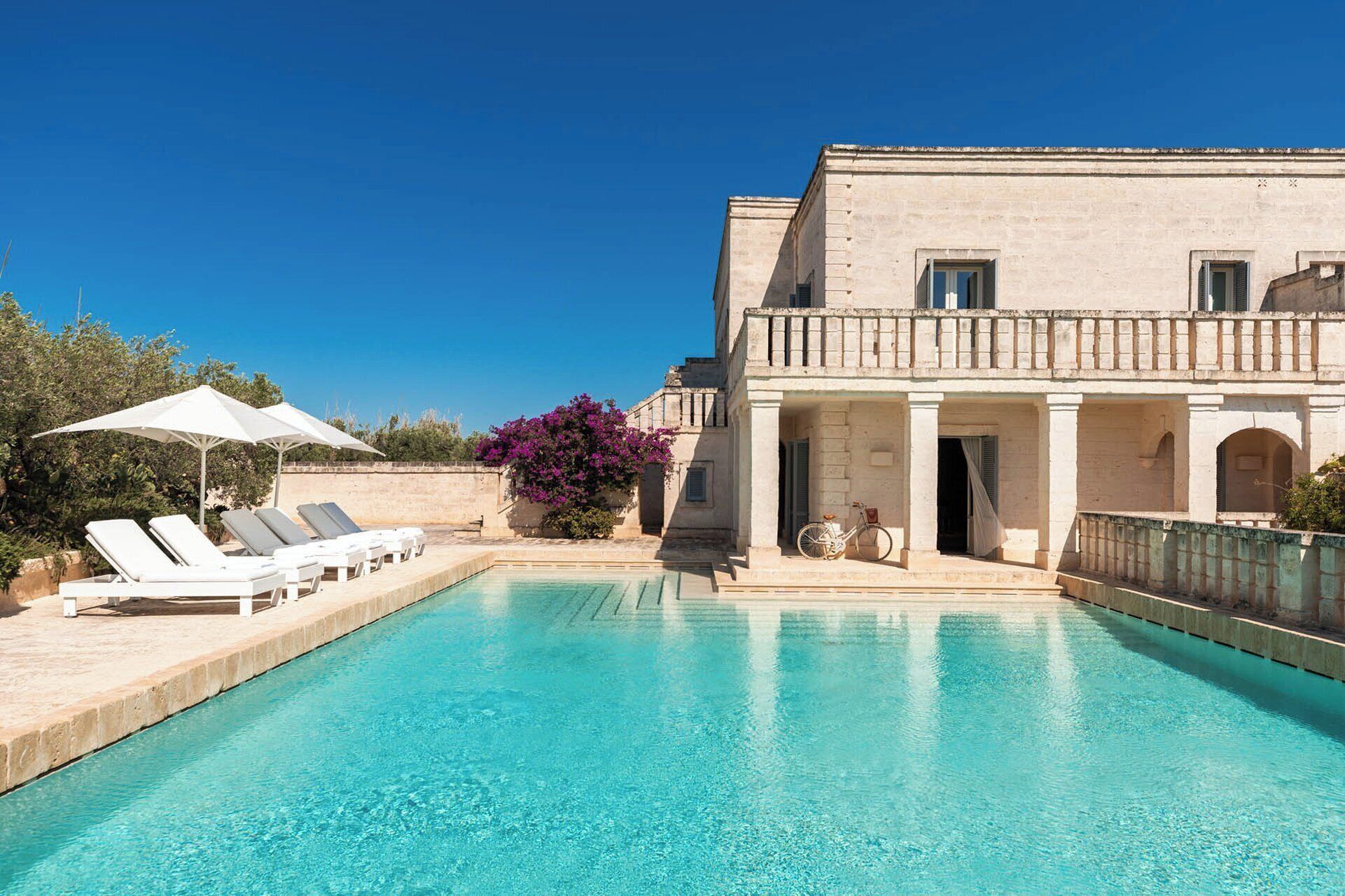 A swimming pool with lounge chairs and umbrellas, a building with a balcony, and a bicycle.
