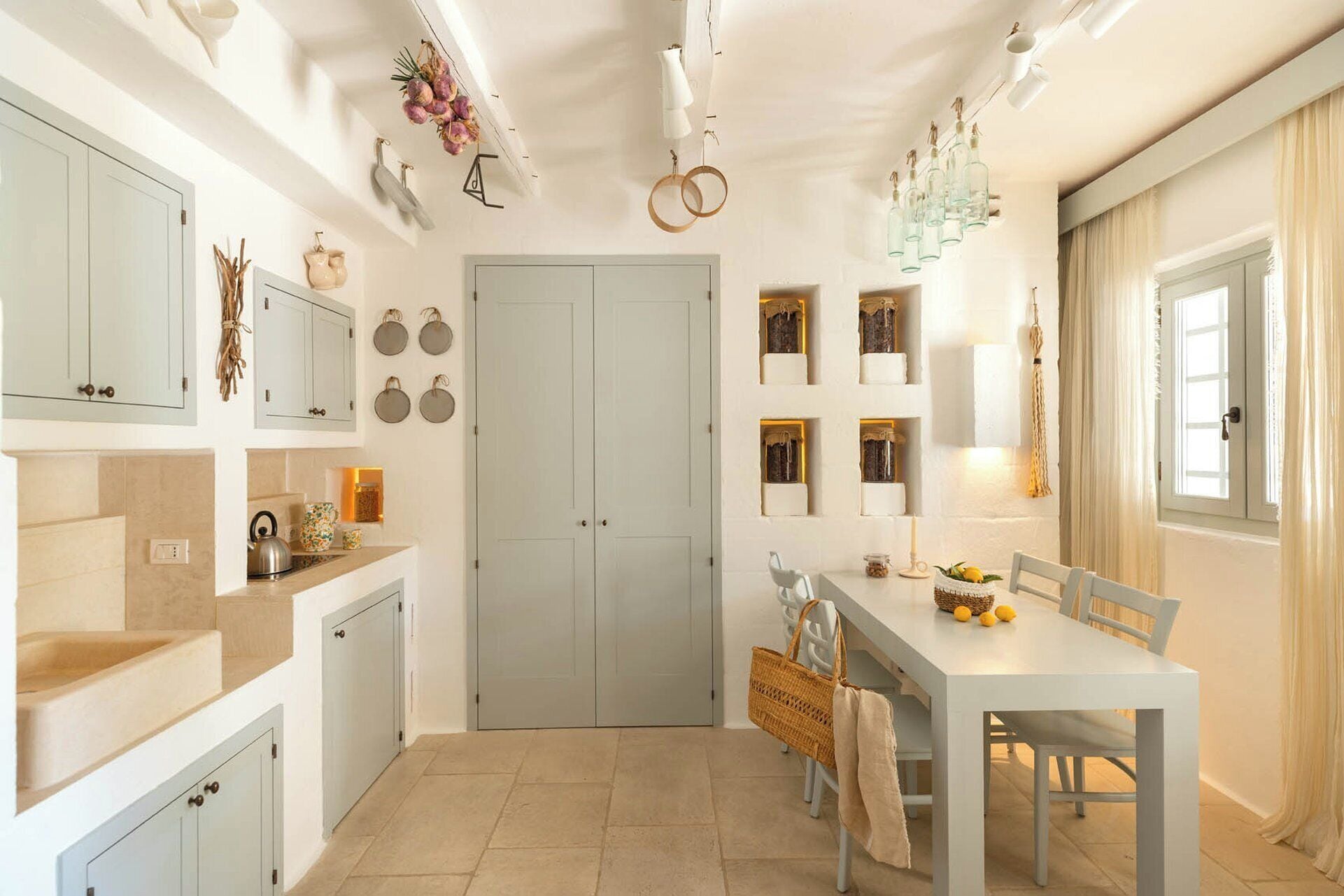 Una cocina con armarios blancos, una mesa de comedor con sillas y un ventanal con cortinas.