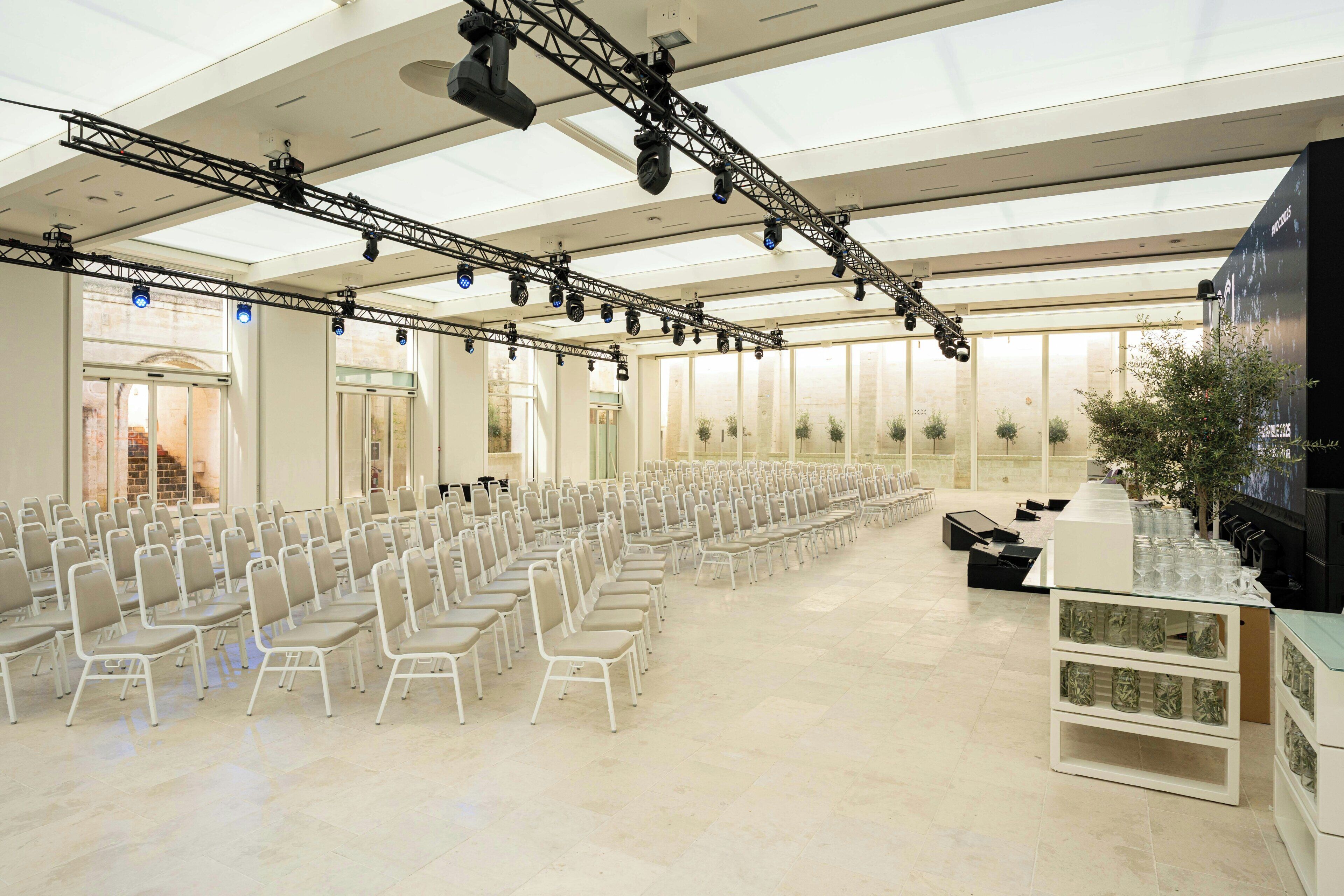 Un ampio spazio interno con file di sedie, un palco con attrezzature luminose e un grande schermo.