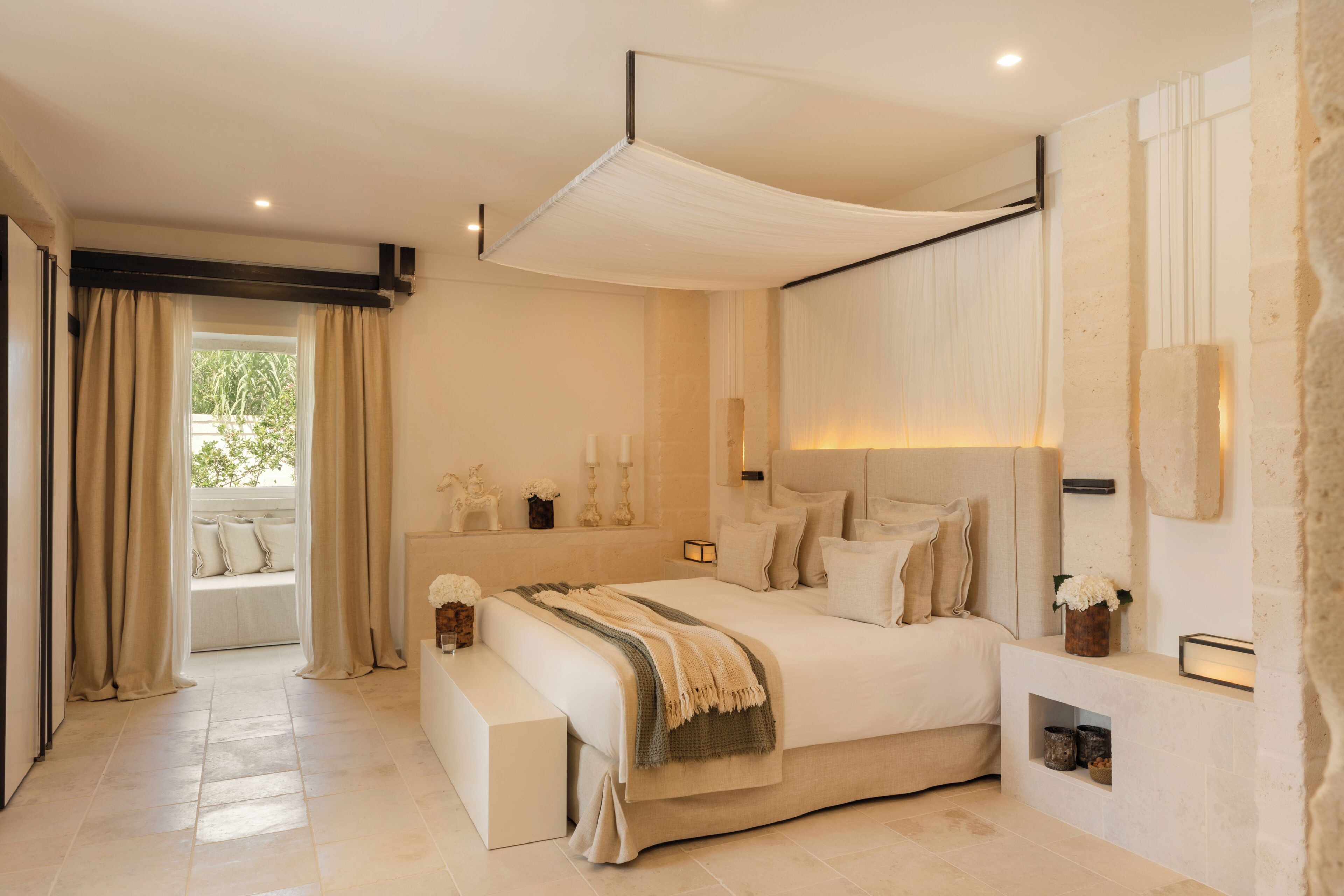 A bedroom with a large bed, a bench, a bedside table, a wall-mounted light, and a window with curtains.