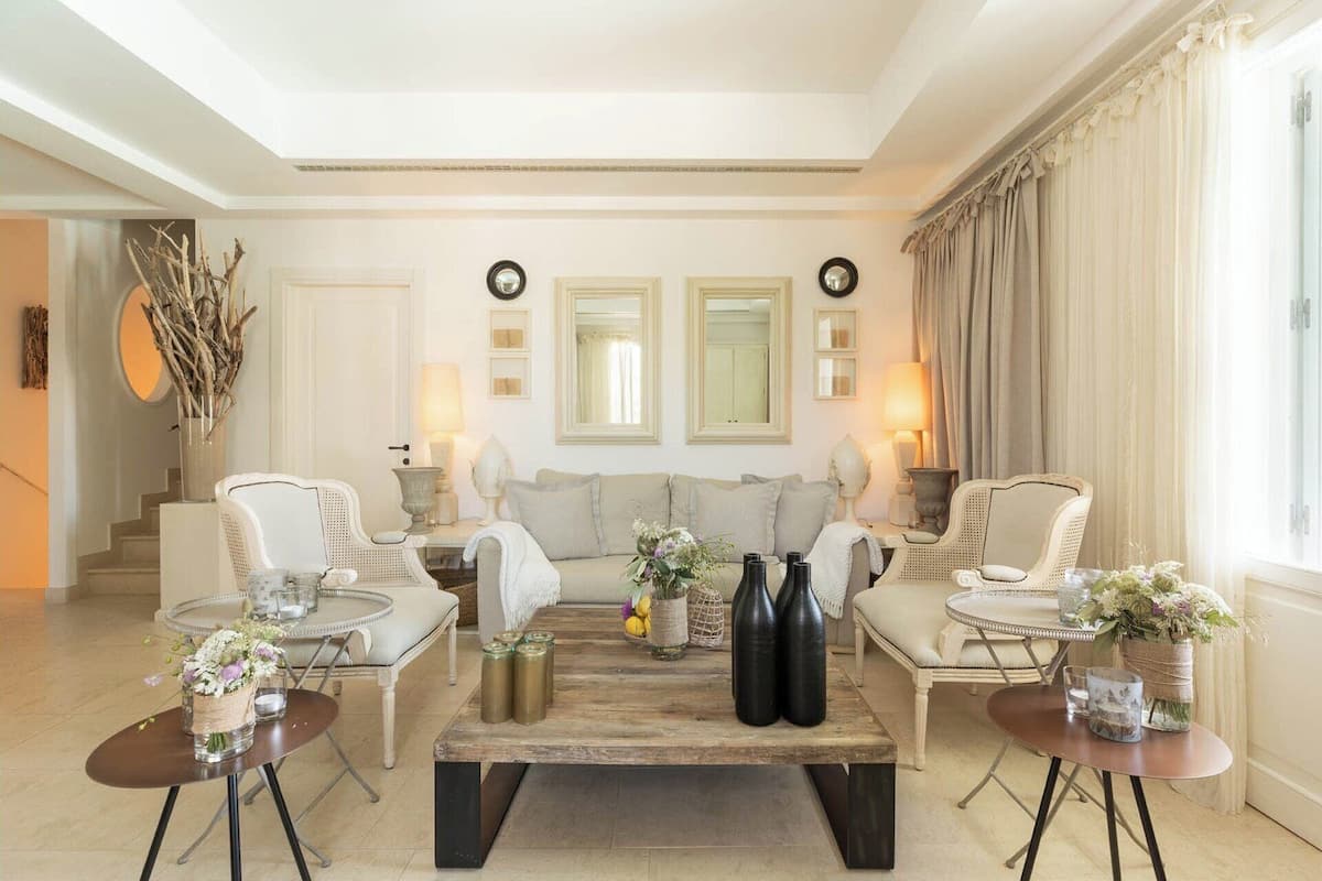A spacious living room with a central coffee table, two armchairs, a sofa, and a dining table with chairs. There are two wall-mounted clocks and a large window with curtains.
