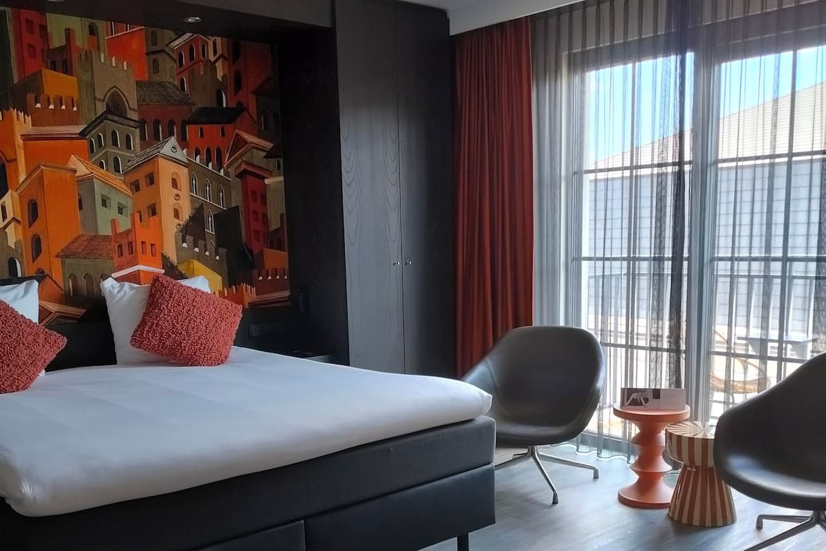 A modern hotel room with a large bed, a cityscape mural, a large window with curtains, and two chairs.