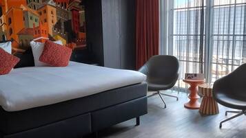 Una habitació d'hotel moderna amb un llit gran, un mural amb un paisatge urbà, una finestra ampla amb cortines i dues cadires.