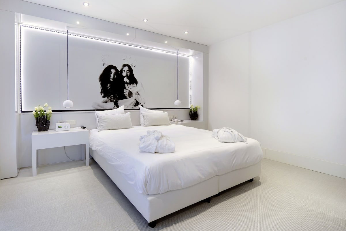 A modern hotel room with a large bed, a bedside table with a radio, a mirror with LED lighting, and a wall-mounted artwork.