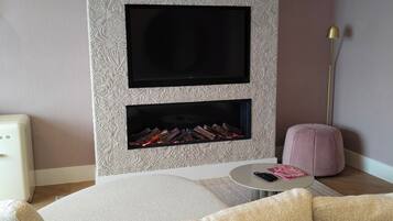Ein modernes Wohnzimmer mit einem flachbildfernseher an einer strukturierten Wand, einem kleinen runden Tisch und einem rosa Pouf.