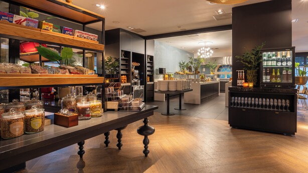 Un moderno vestíbulo de hotel con una larga recepción, estantes con aperitivos, una zona de bar con botellas y un área de estar con vistas.