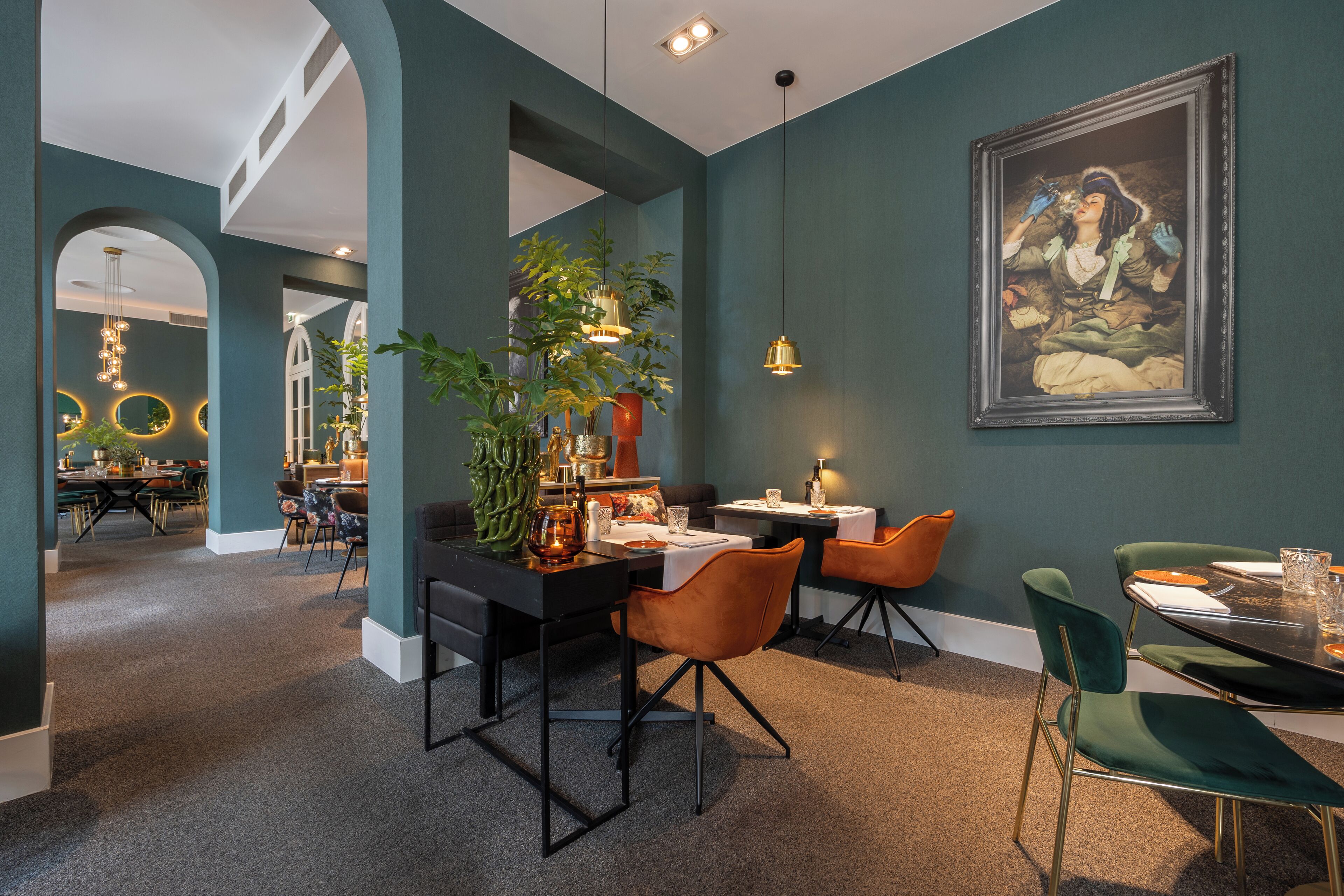 A modern dining area with a black table, orange chairs, and a green chair. There is a potted plant on the table and a painting on the wall.