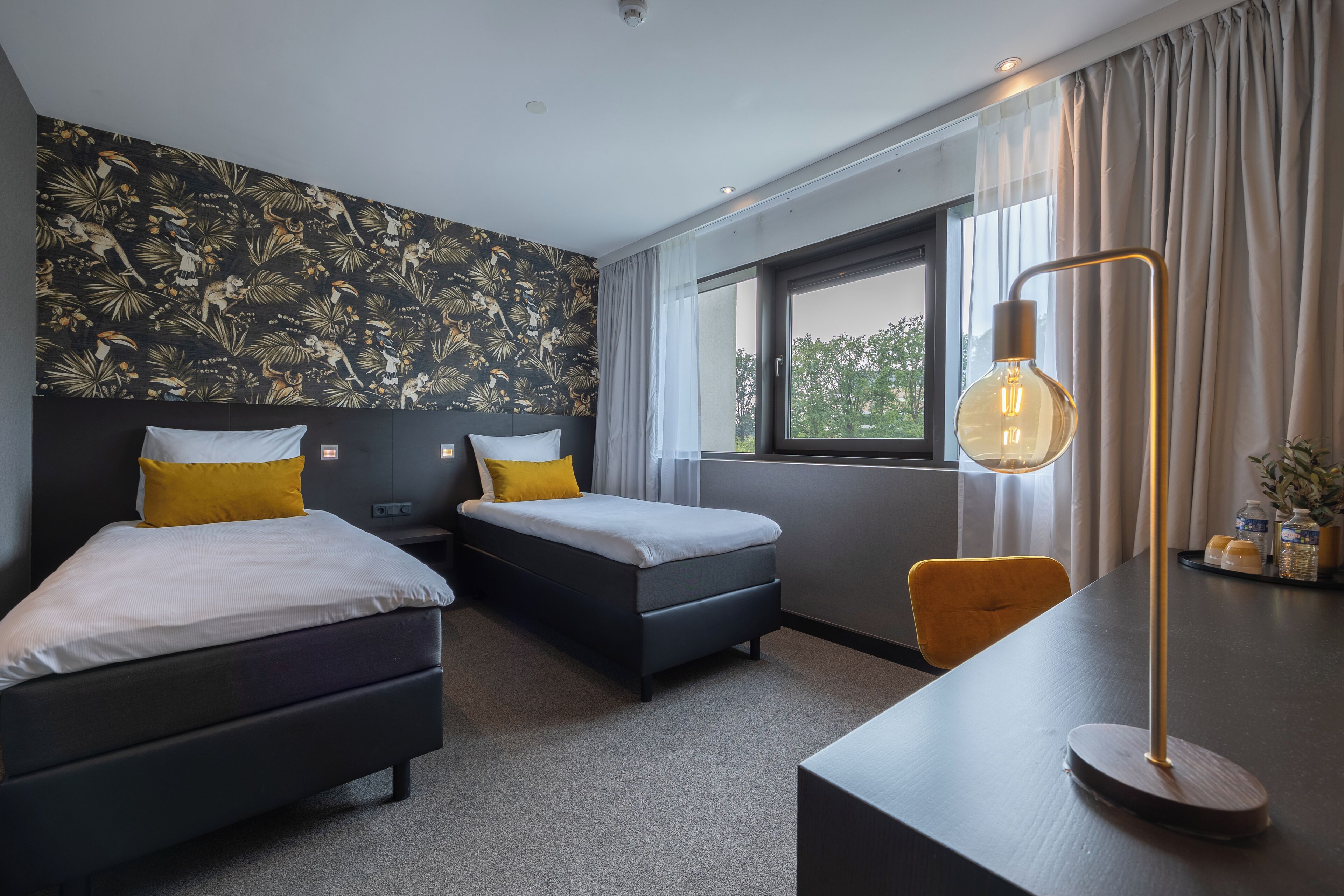 A modern hotel room with two beds, a desk, a chair, a large window, and a decorative wall.