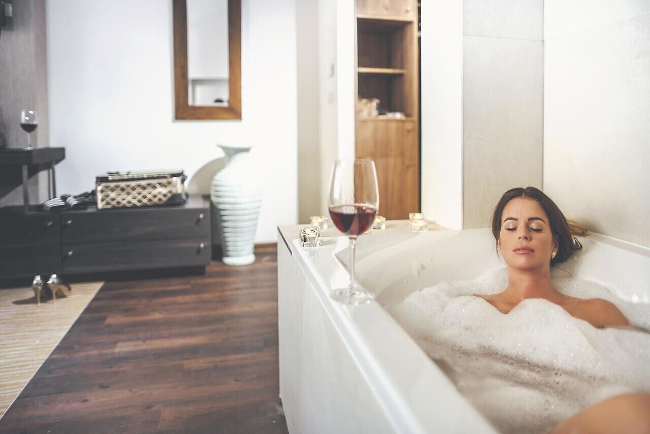A person relaxing in a bathtub with a glass of red wine on the side.