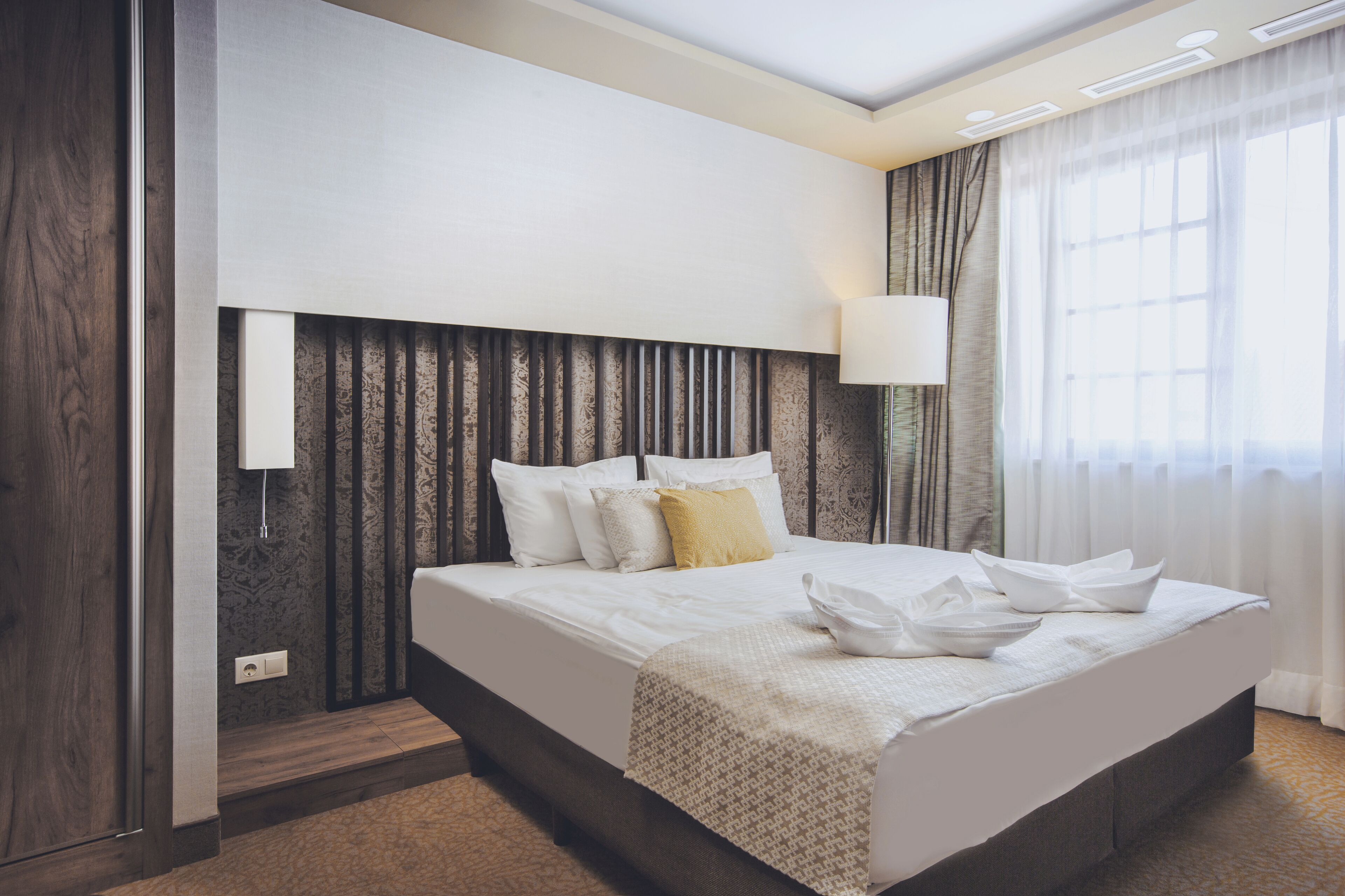 Ett modernt hotellrum med en stor säng, en sänglampa och utsikt över omgivningarna genom tunna gardiner.