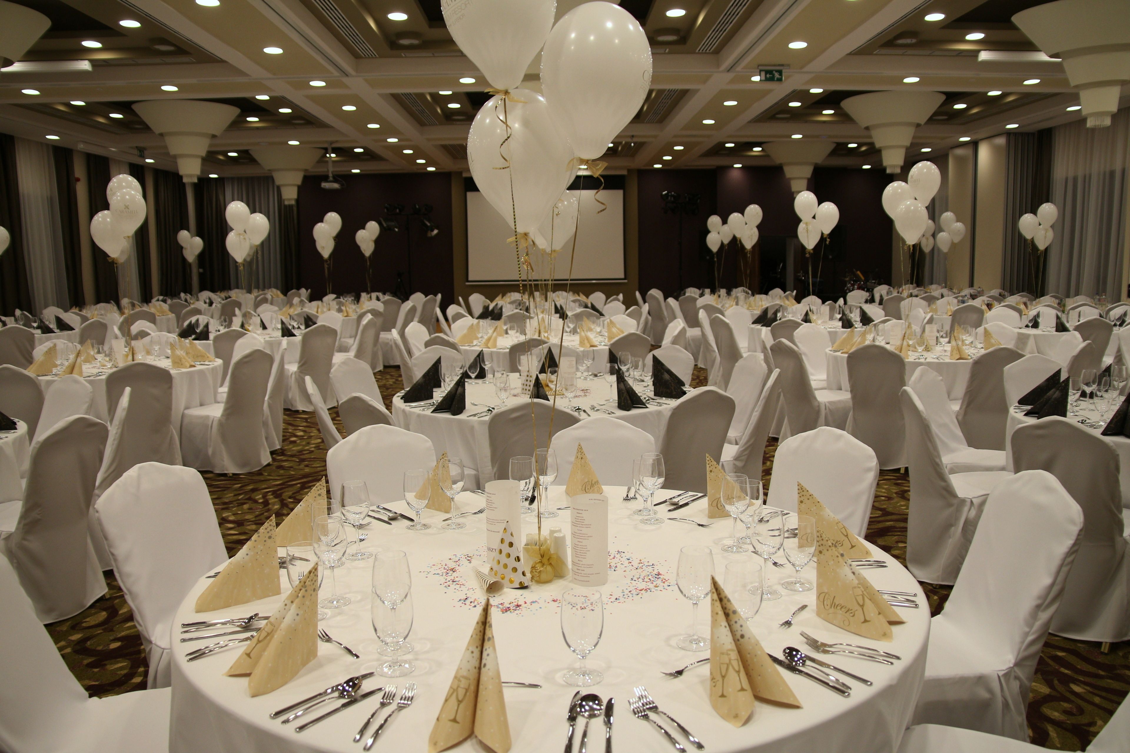 A banquet hall with round tables set for a formal event, featuring white tablecloths, gold centerpieces, and white chairs.