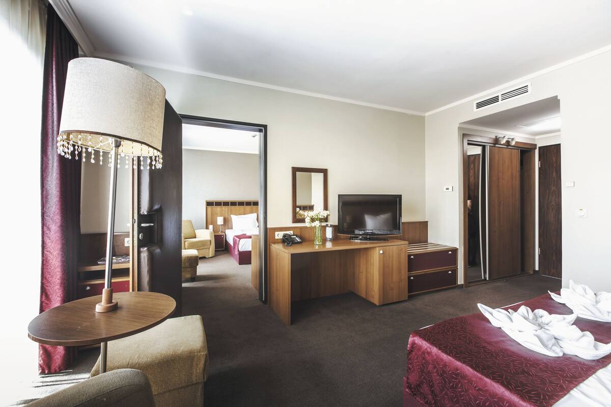 Ein Hotelzimmer mit einem Holztisch, einem Fernseher, einem Bett mit roter Bettdecke, einem runden Tisch mit Lampe und einem Spiegel an der Wand.