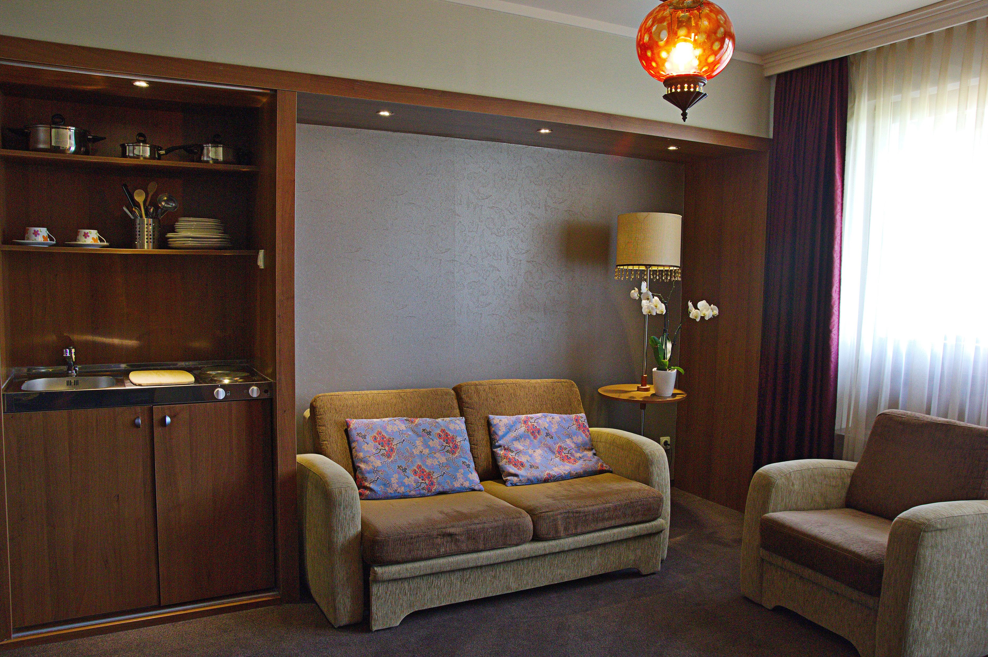 A compact living space with a kitchenette, a sofa with floral cushions, a side table with a lamp, and a view of the outdoors through a window with curtains.