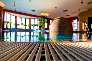 An indoor swimming pool with a tiled wall, potted plants, and a ladder leading to a higher level. - Caramell Premium Resort (Buk)