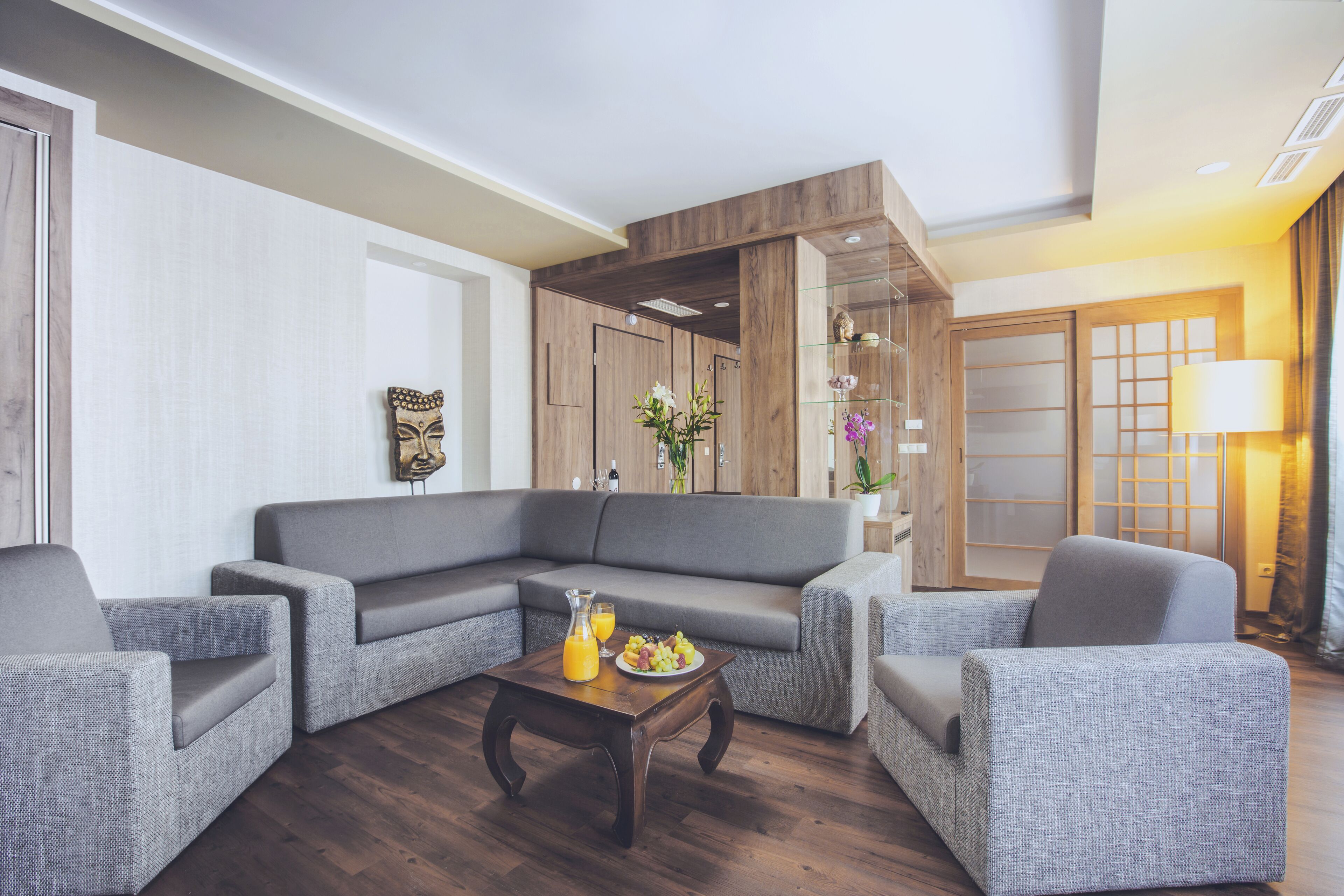 A modern living room with a grey sectional sofa, two armchairs, a wooden coffee table with fruit and a glass of orange juice, and a decorative mask on the wall.