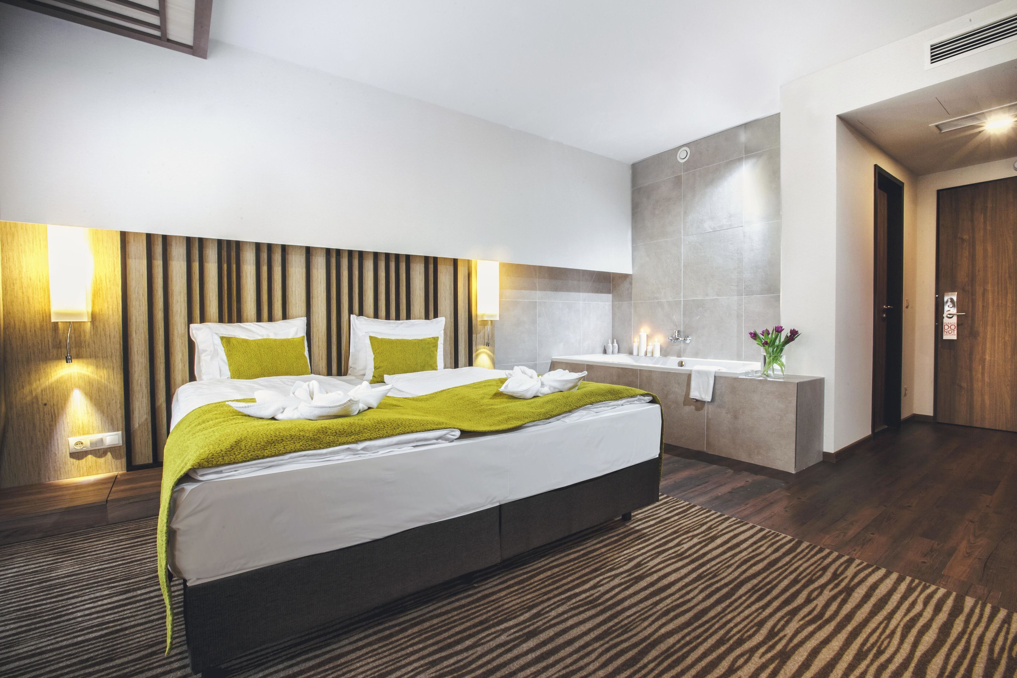 A modern hotel room with a large bed, a bedside table, a built-in vanity with a mirror, and a shower area.