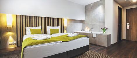 A modern hotel room with a large bed, a bedside table, a built-in vanity with a mirror, and a shower area.