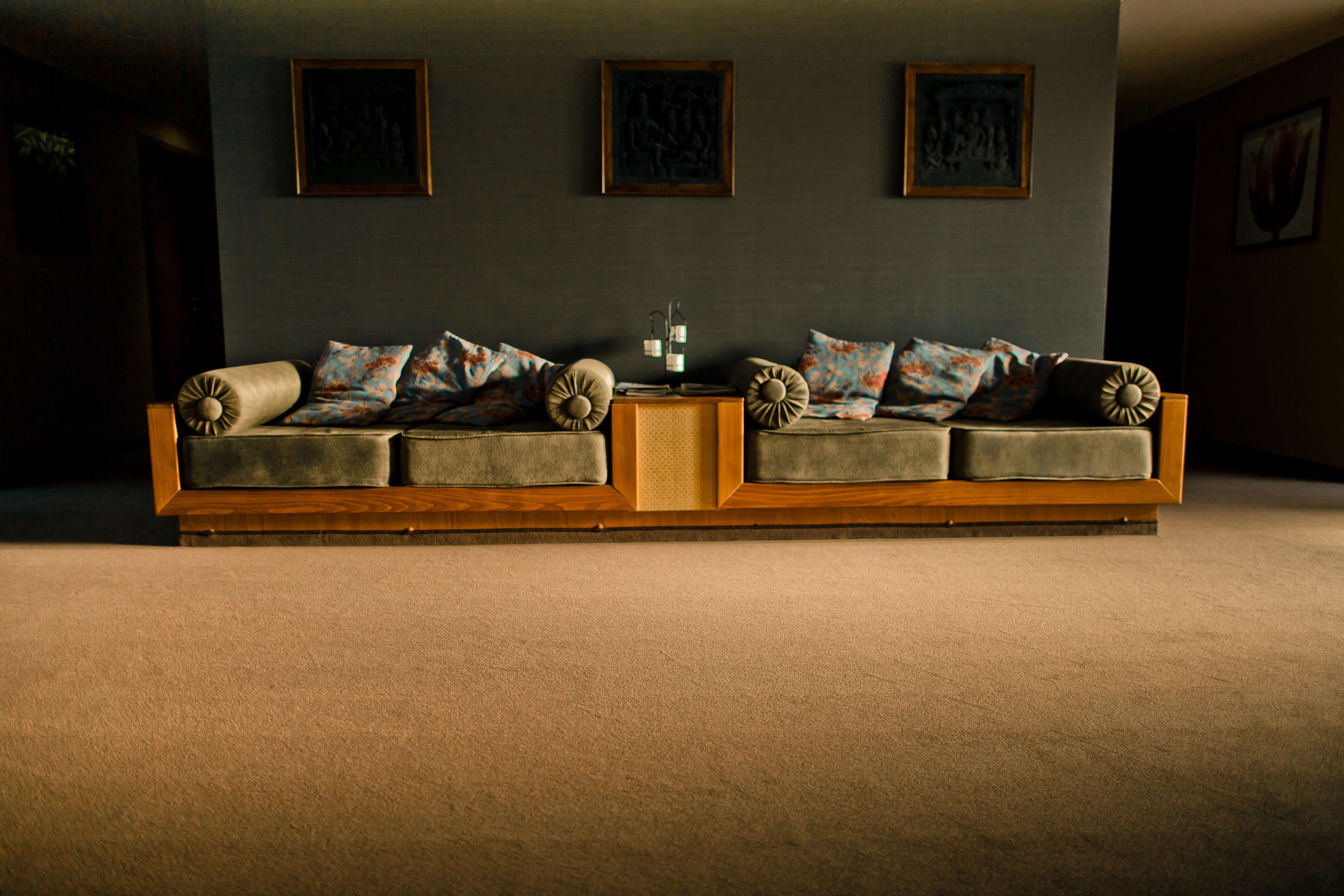A long, wooden sofa with multiple cushions against a dark wall with three framed artworks.