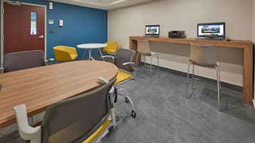 A conference room with a wooden table, yellow and gray chairs, a white round table, and two computer monitors on a wooden desk.