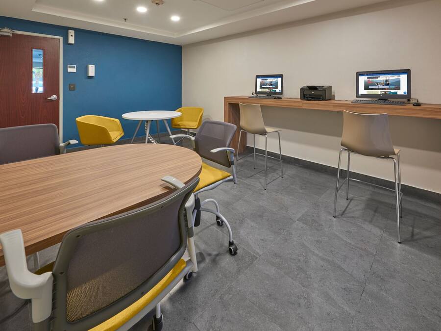 A conference room with a wooden table, yellow and gray chairs, a white round table, and two computer monitors on a wooden desk.