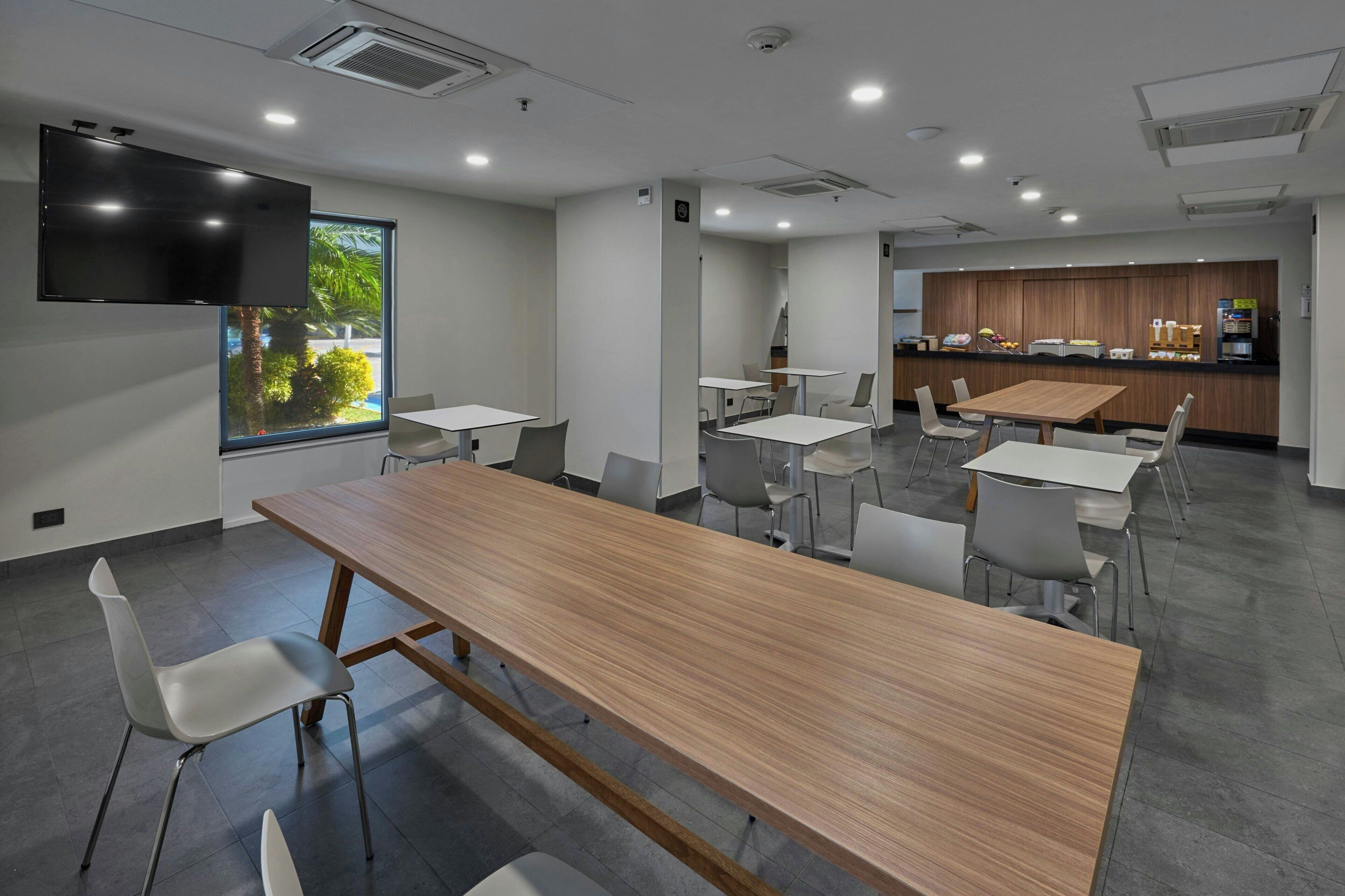 A modern meeting room with a long wooden table, several white chairs, a flat-screen TV mounted on the wall, and a kitchen area in the background.
