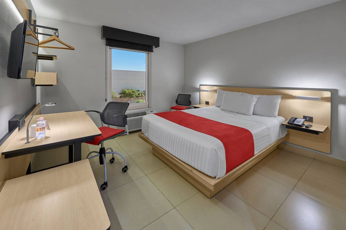 Quarto de hotel com cama, escrivaninha, cadeira e televisão na parede.