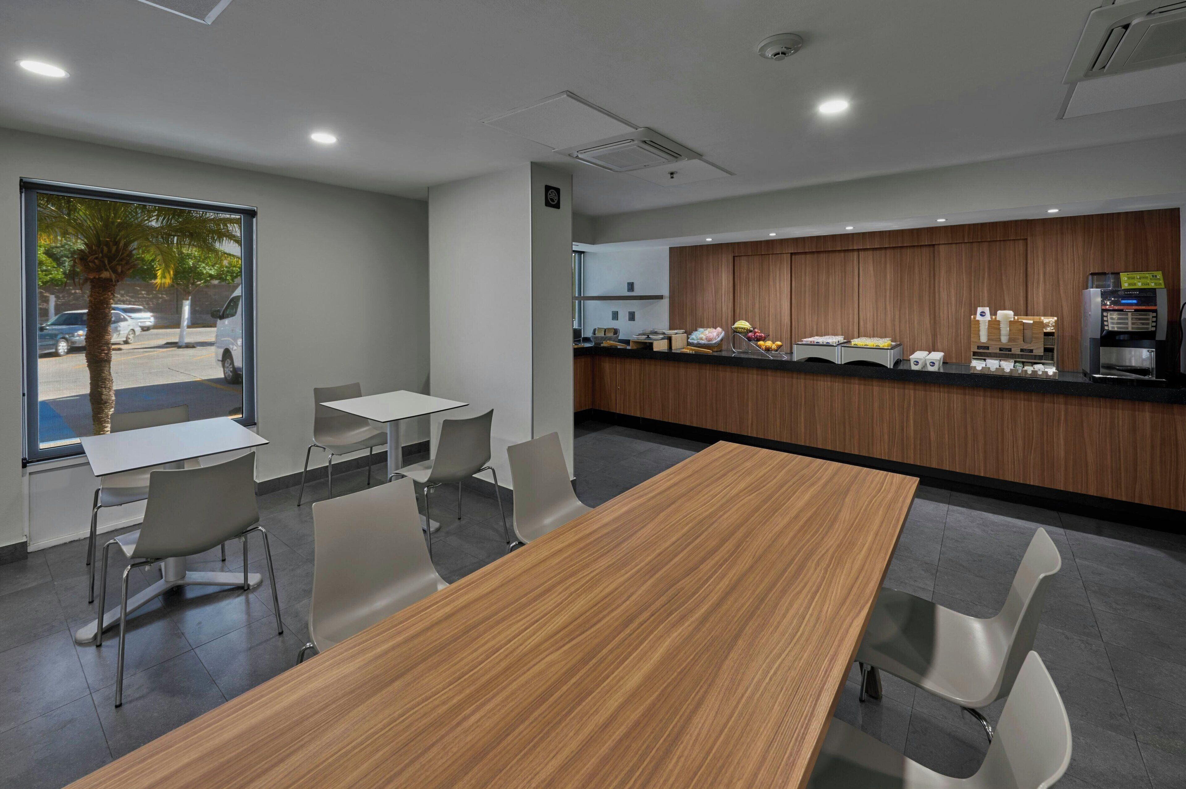 A modern dining area with a wooden table and chairs, a counter with a coffee machine, and a view of parked cars outside.