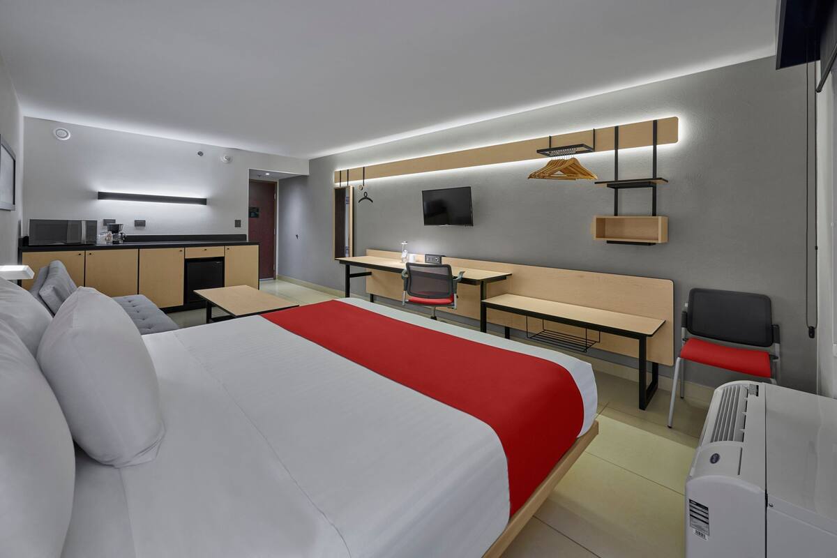 Quarto de hotel moderno com uma cama grande, uma escrivaninha, uma cadeira, uma televisão e uma cozinha compacta.