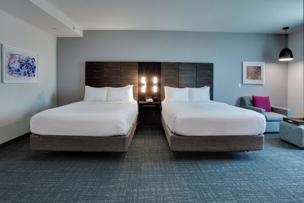 Ein Hotelzimmer mit zwei Betten, einem dunklen Kopfteil, einem grauen Teppich, einem grauen Sessel und einem kleinen Tisch.