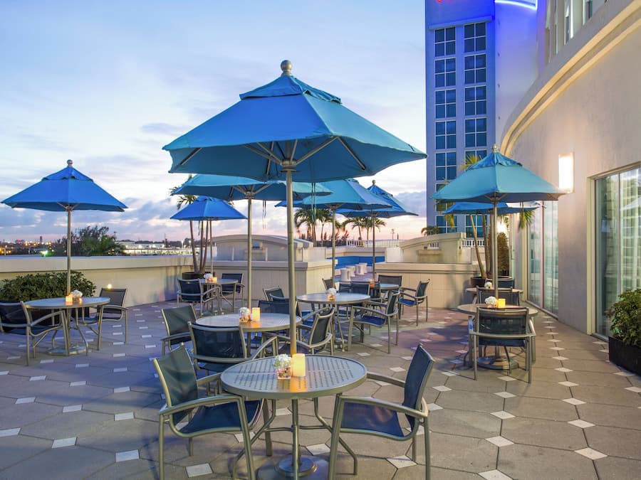 An outdoor seating area with round tables and chairs, blue umbrellas, and a view of the city skyline.