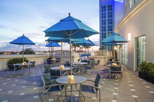 An outdoor seating area with round tables and chairs, blue umbrellas, and a view of the city skyline.