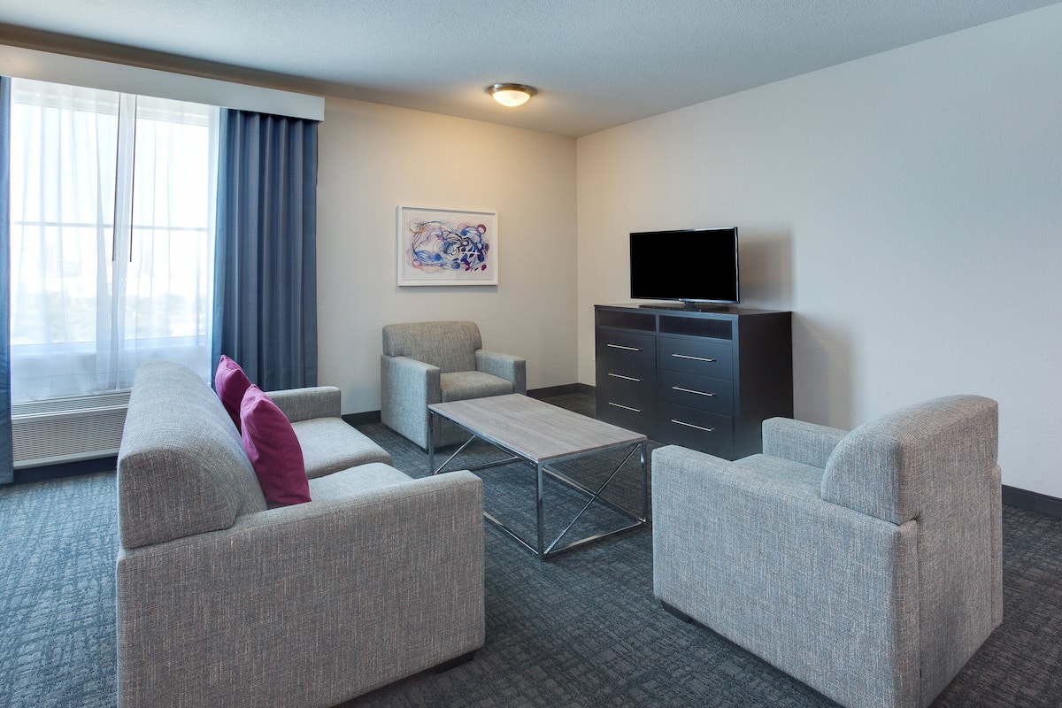 Ein Hotelzimmer mit grauem Sofa, einem kleinen Sessel, einem Couchtisch, einem Flachbildfernseher und einem Gemälde an der Wand.