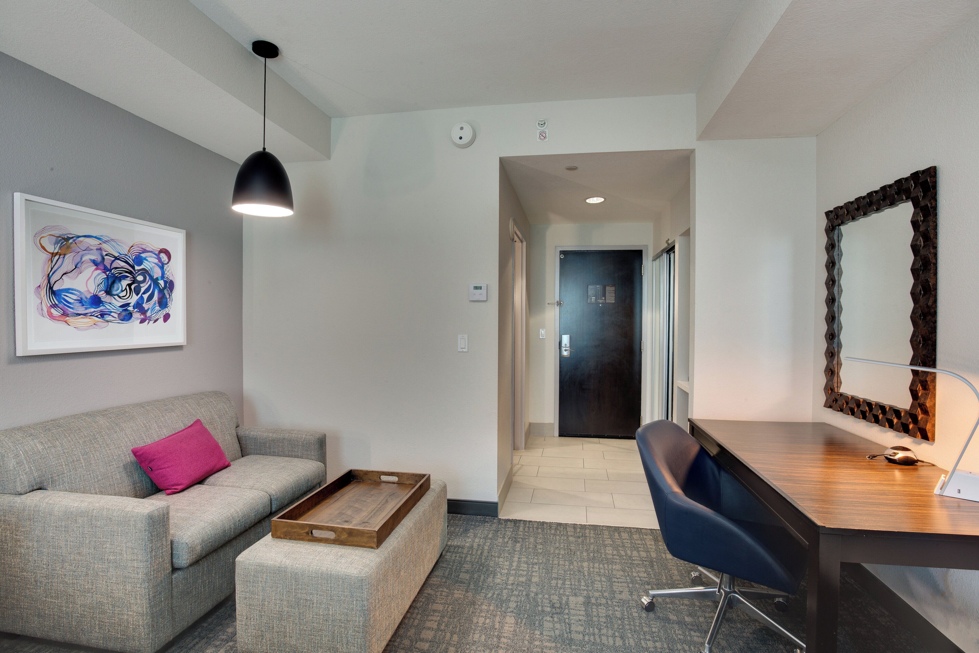 A modern hotel room with a grey sofa, a wooden side table, a desk with a chair, a large mirror, and a framed abstract painting.
