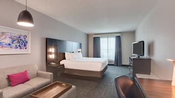 A hotel room with a bed, a desk, a chair, a sofa, a TV, and a painting on the wall.