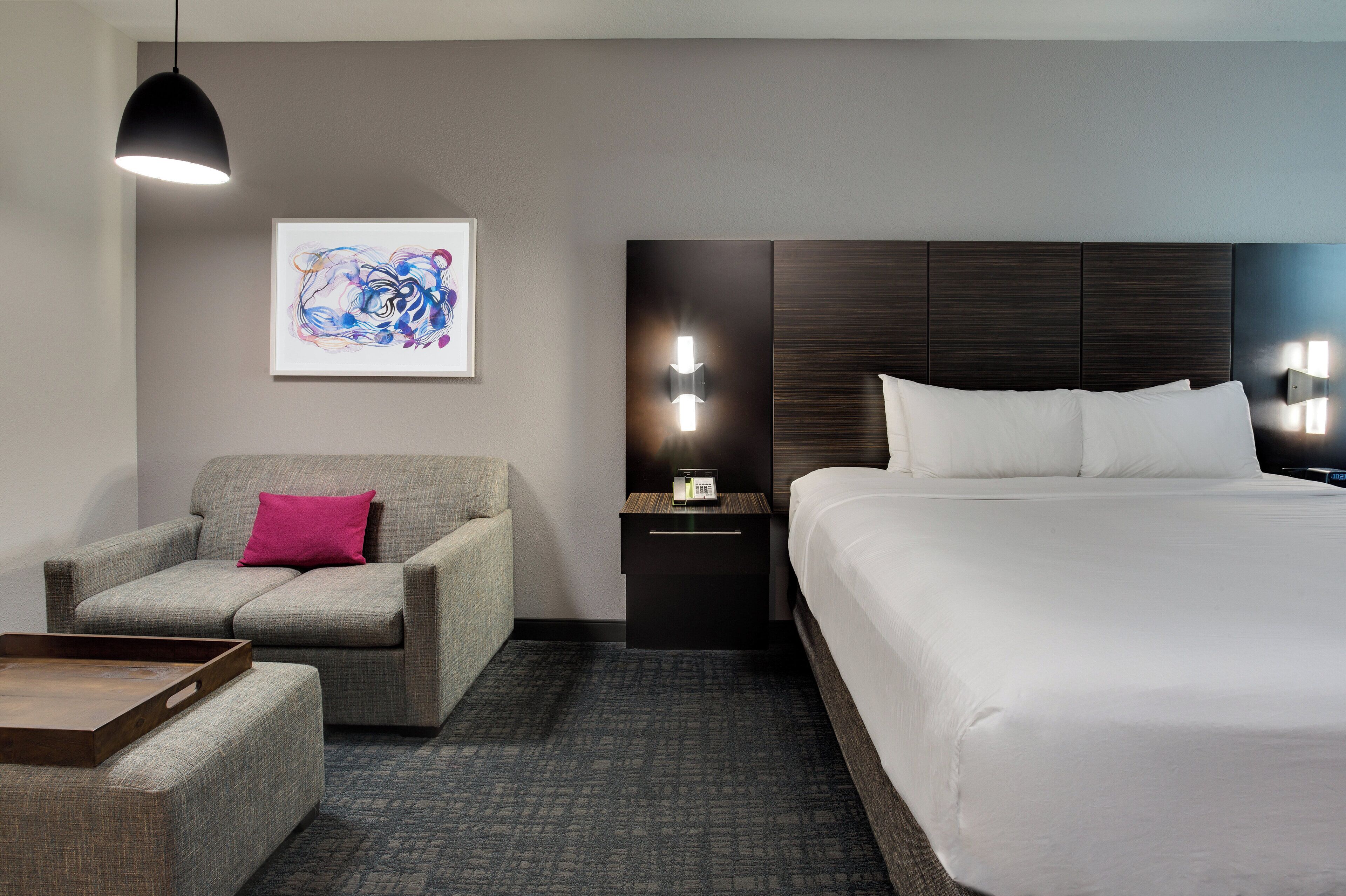 Una habitación de hotel con cama, una silla, una mesita de noche, una lámpara y un cuadro en la pared.