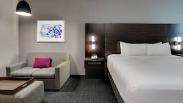 Una habitación de hotel con cama, una silla, una mesita de noche, una lámpara y un cuadro en la pared.