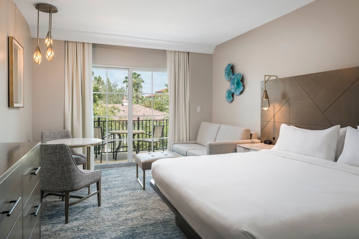 Una camera d'albergo con un letto grande, una scrivania, una sedia, un divano e un balcone con vista sulle palme.