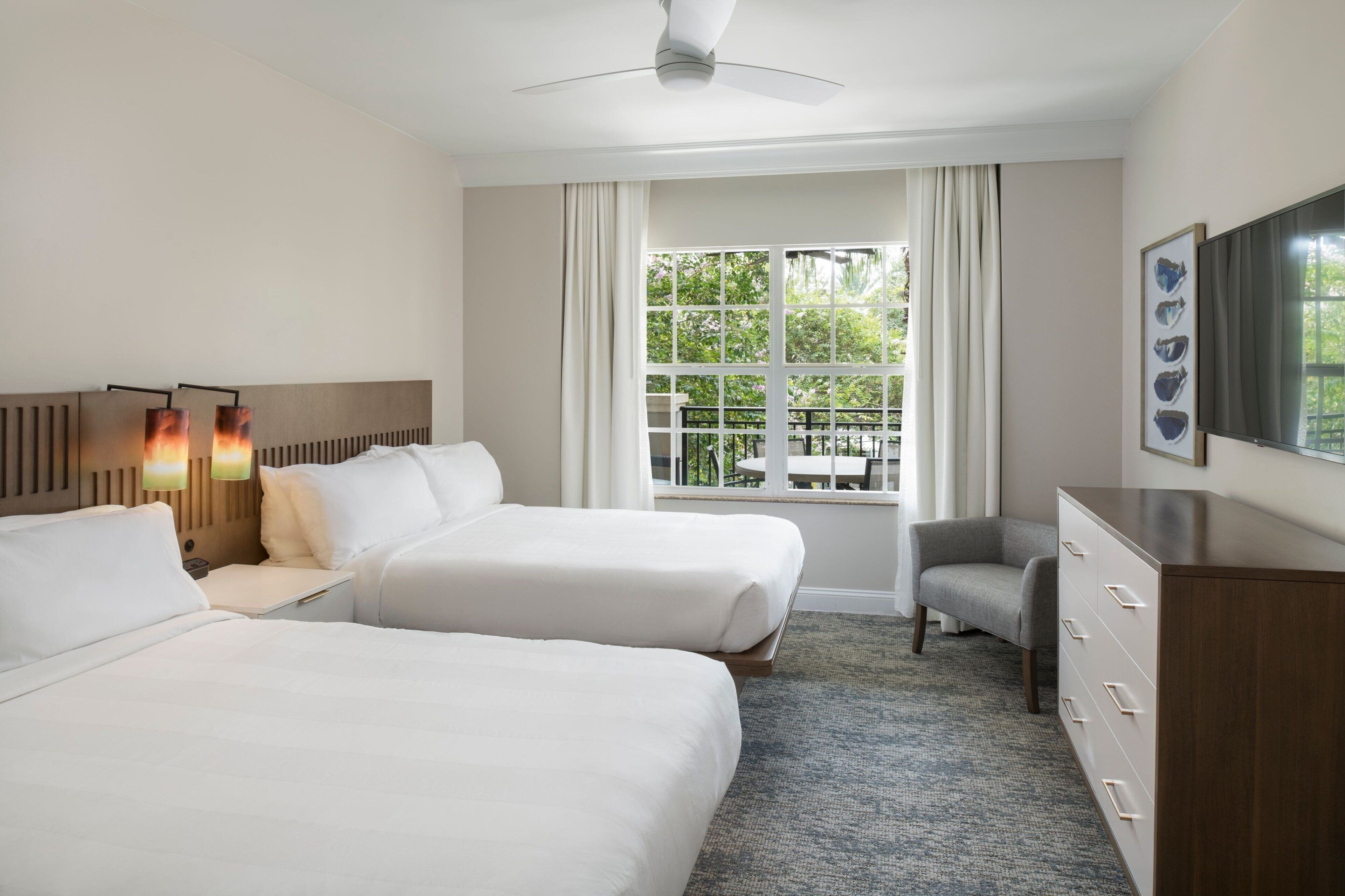 A hotel room with two beds, a desk, a chair, a dresser, a ceiling fan, and a window with a view of greenery.