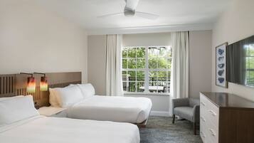 A hotel room with two beds, a desk, a chair, a dresser, a ceiling fan, and a window with a view of greenery.