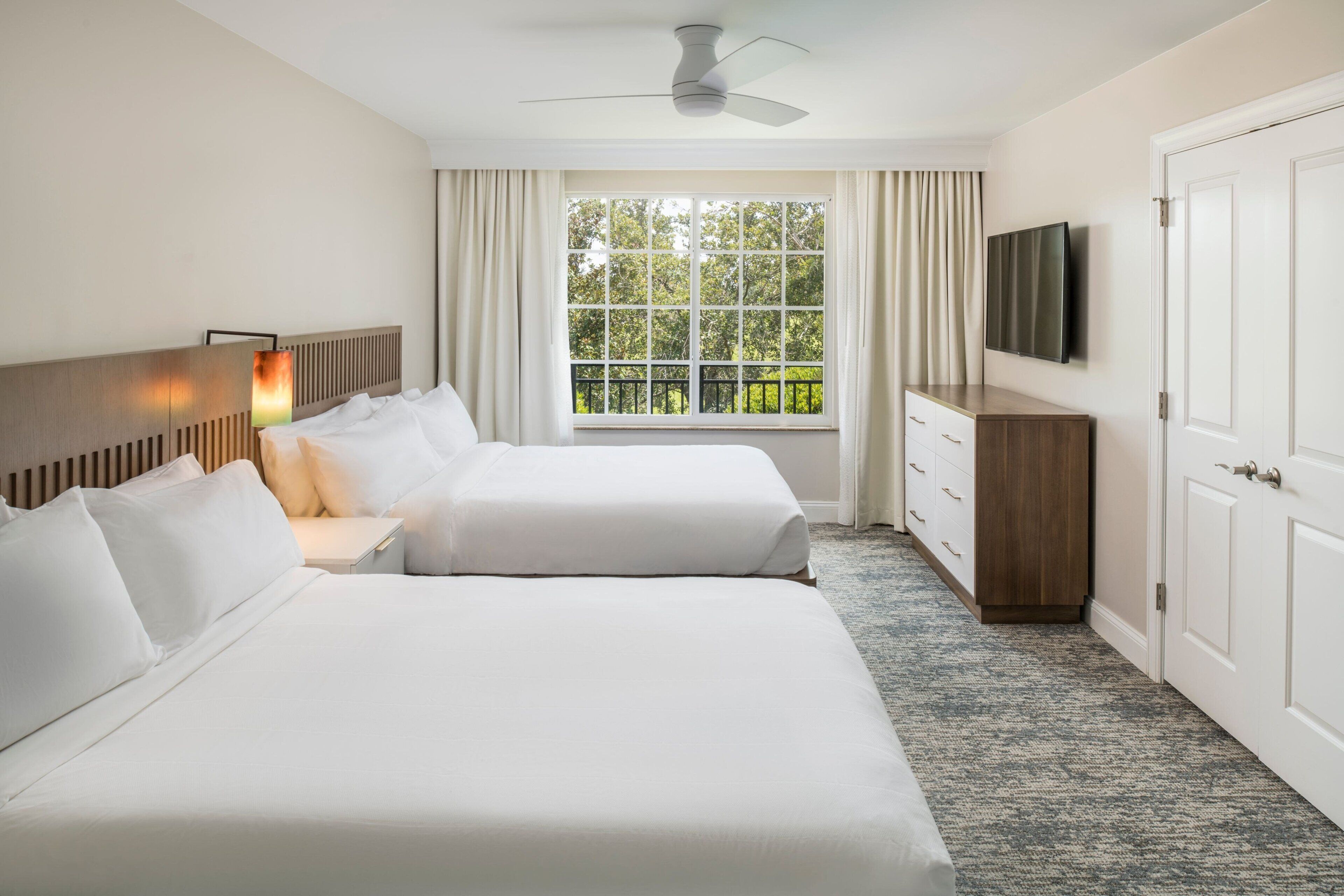 Habitación de hotel con dos camas, un televisor, ventilador de techo, ventana con vista a la vegetación y cómoda de madera.