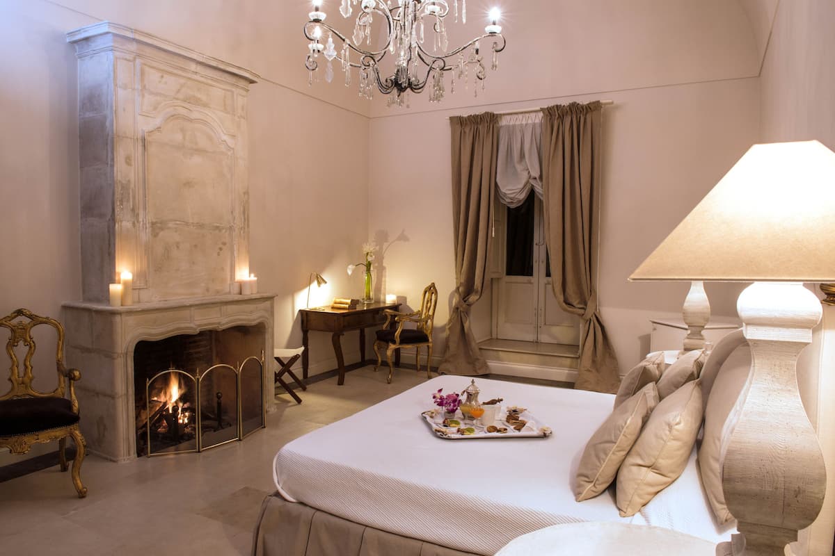 Quarto com lareira, candelabro, cama com almofadas, bandeja com comida e escrivaninha com cadeira.