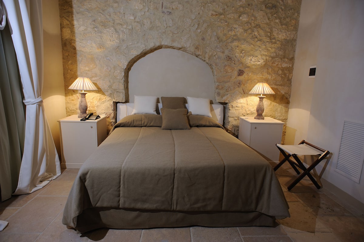 Quarto com parede de pedra, cama com cabeceira, duas mesas de cabeceira com luminárias e uma cadeira.